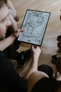 Über Mich Coach presents a basketball strategy on clipboard during indoor team huddle.