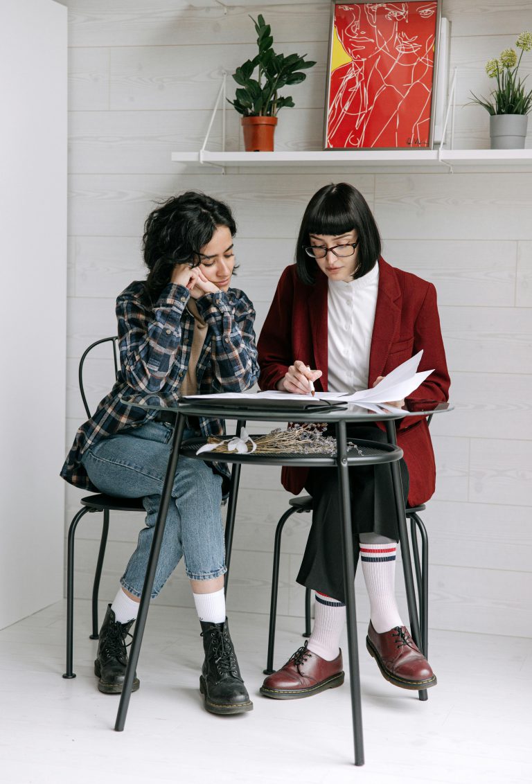 Two women discuss ideas at a table in a cozy indoor setting, surrounded by art and plants.